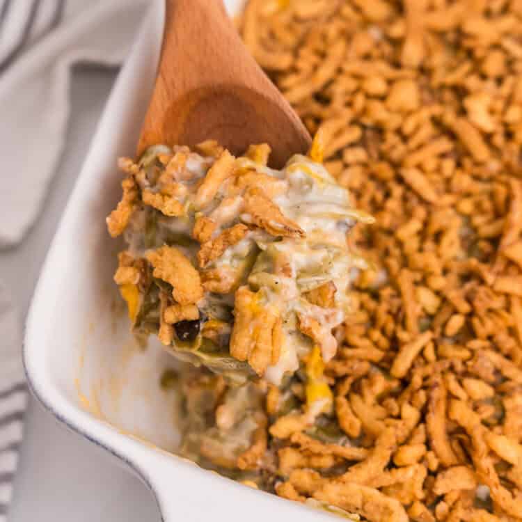 A wooden spoon scooping out a serving of creamy green bean casserole topped with crispy onions from the baking dish.