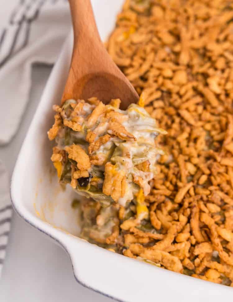 A wooden spoon scooping out a serving of creamy green bean casserole topped with crispy onions from the baking dish.