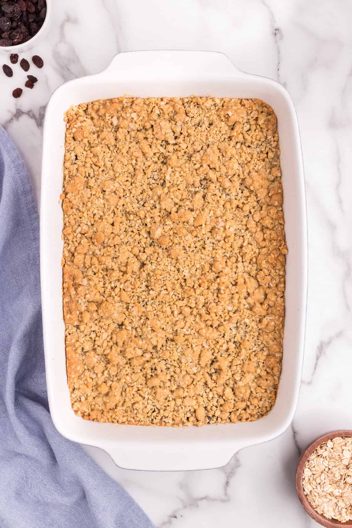 A pan of baked sour cream raisin bars with a golden brown oat crumble topping, cooling in a white baking dish. A few oats and raisins are scattered nearby for styling.