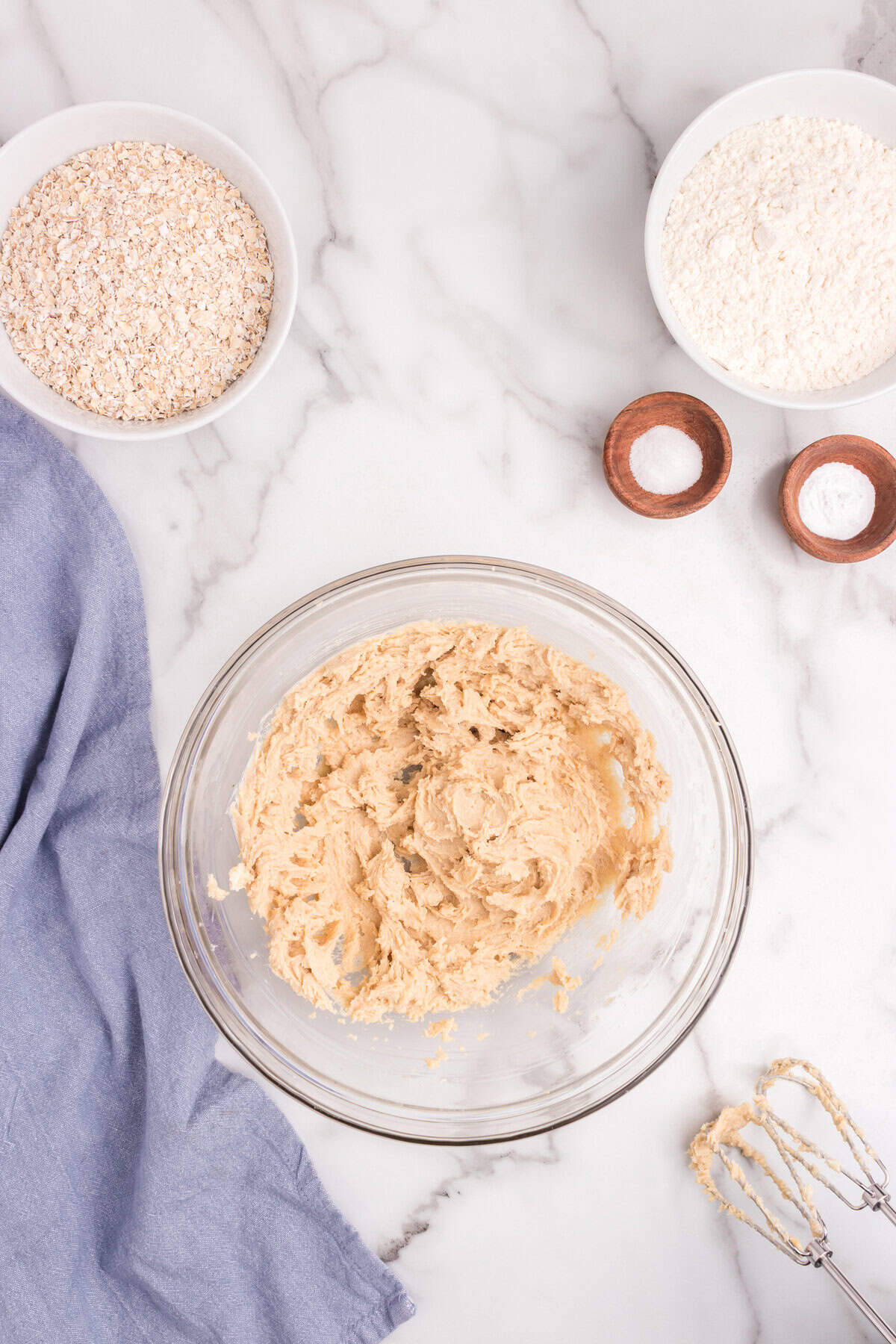 A glass mixing bowl with creamed butter and sugar mixture on a marble counter. Around it are bowls with oats, flour, baking powder, and baking soda, along with a hand mixer and a blue linen cloth.