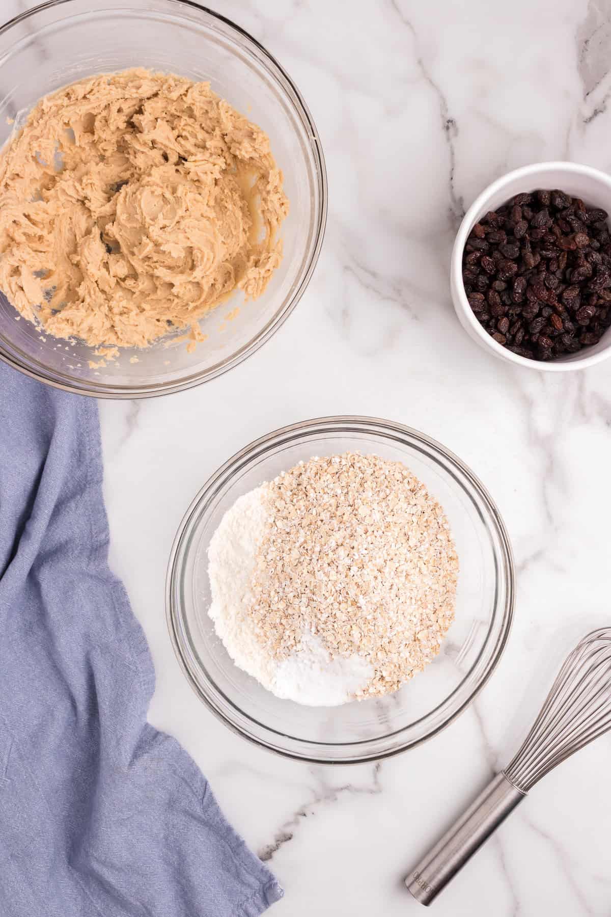 A bowl with creamed butter mixture next to another bowl filled with oats and flour mixture. A small bowl of raisins and a whisk are also on the counter.