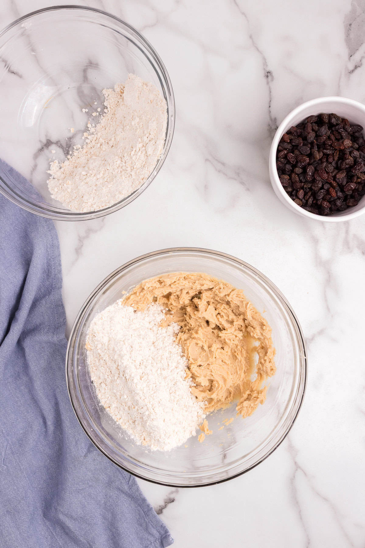 The butter mixture in a mixing bowl with dry ingredients (flour and oats) partially added in. Another bowl with leftover dry mix and a small bowl of raisins are nearby.