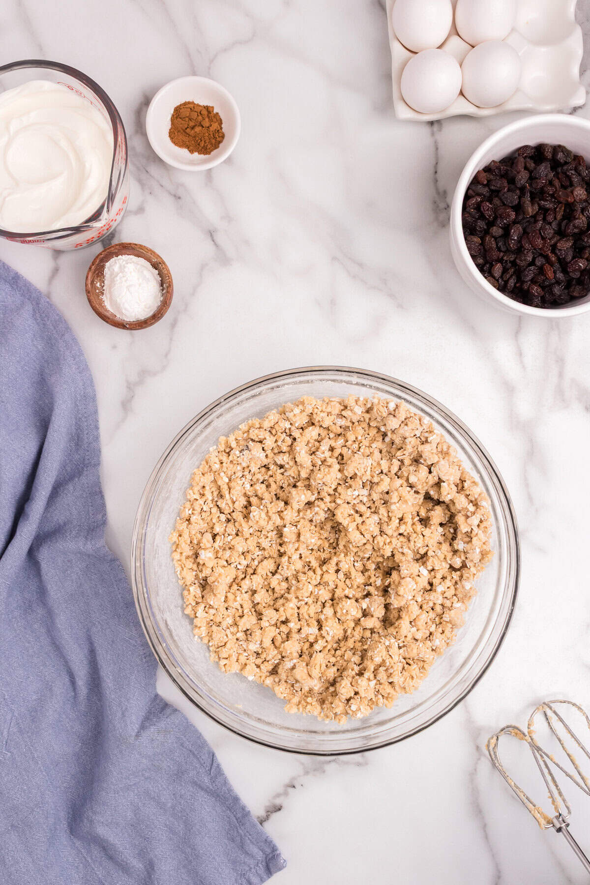 A bowl containing combined oats and flour mixture mixed into the butter mixture, creating a crumbly dough. Ingredients like sour cream, cinnamon, eggs, and raisins are visible around the counter.