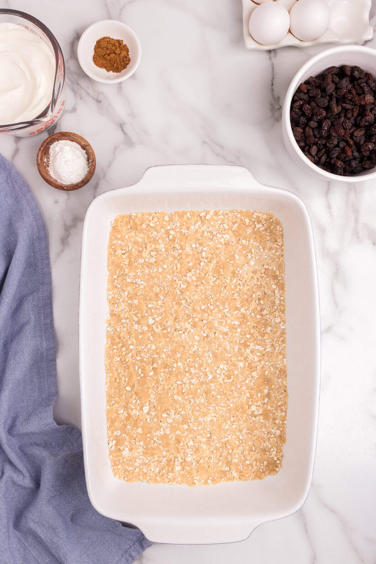 A baking dish with the oat crust mixture evenly pressed into the bottom, ready for the filling. Additional ingredients remain on the side, including sour cream, eggs, cinnamon, and raisins.