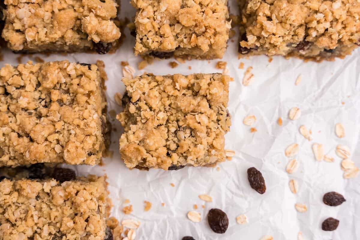 Several sour cream raisin bars arranged on crinkled parchment paper, with loose oats and raisins scattered around, highlighting the crumbly texture and visible raisins in the filling.