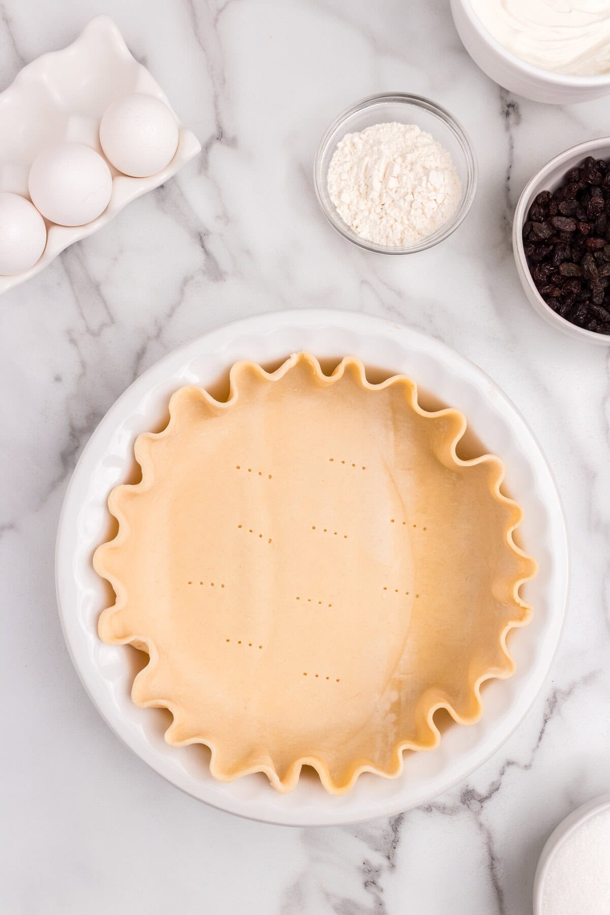 A raw pie crust with fluted edges, docked with fork holes, sits in a white pie dish on a marble surface. Ingredients like eggs, flour, sour cream, and raisins are nearby.