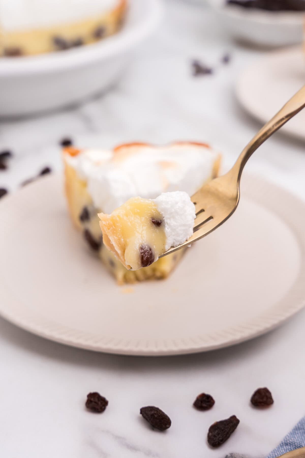 A close-up of a fork holding a bite of sour cream raisin pie with creamy custard, raisins, and fluffy meringue topping, with the rest of the slice on a plate in the background.