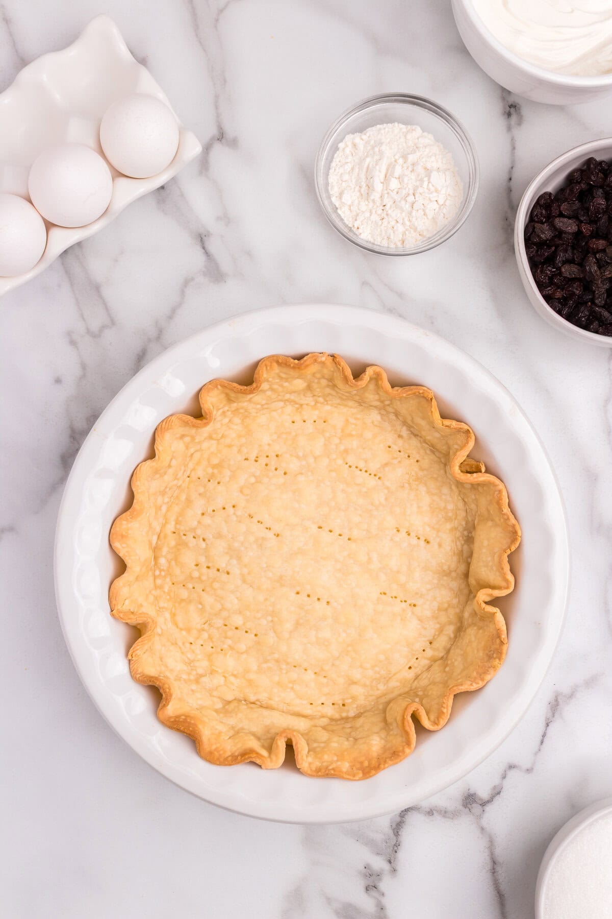 A golden-brown pie crust with fork holes is fully baked in the same white dish, sitting on a marble surface with baking ingredients around.