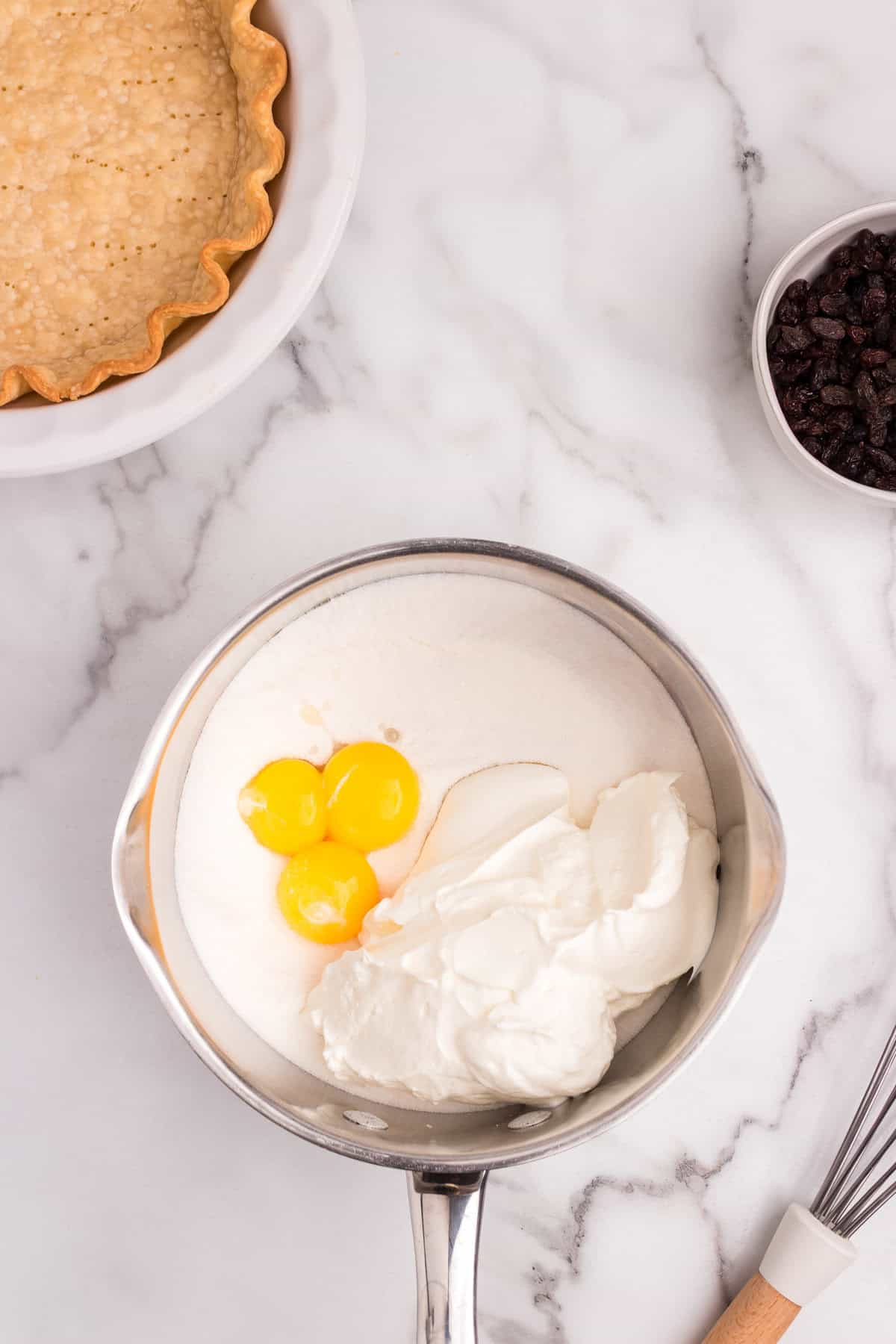 A metal saucepan contains three egg yolks, granulated sugar, and a large scoop of sour cream, ready to be mixed.