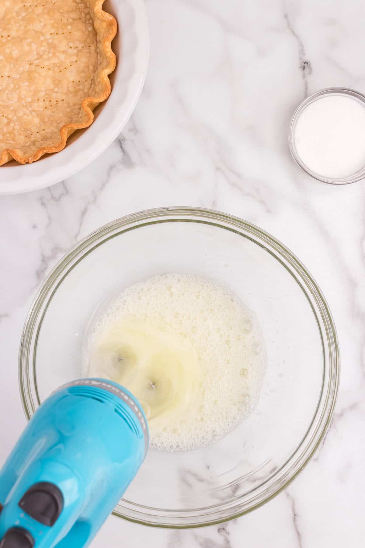 A blue hand mixer is whipping egg whites in a clear glass bowl until frothy.