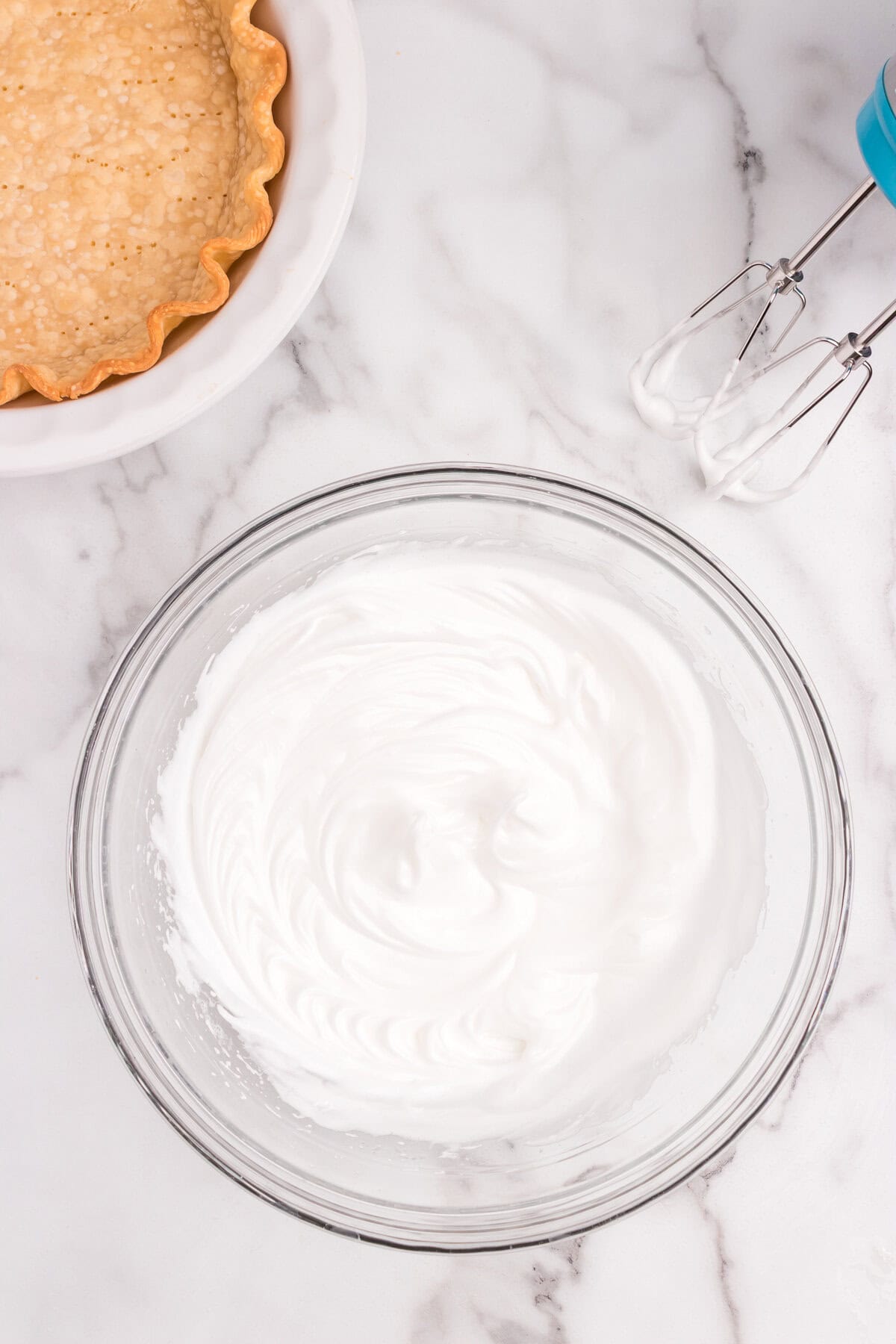 A bowl of glossy, stiffly whipped egg whites sits beside a pie crust and hand mixer.
