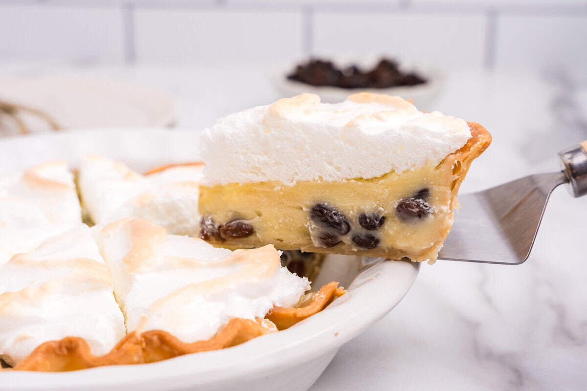 A pie server lifting a slice of sour cream raisin pie from a white dish, showing layers of golden crust, creamy raisin custard, and browned meringue topping.