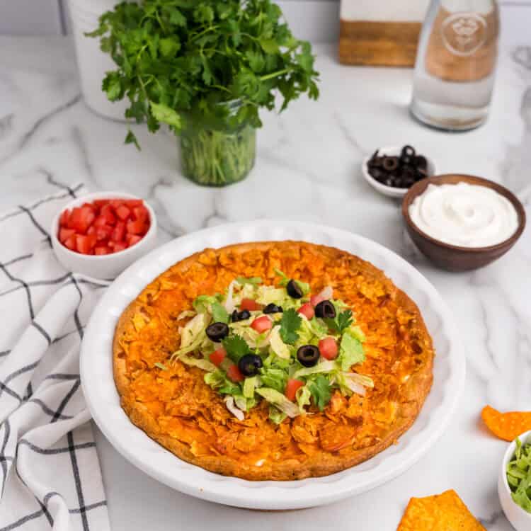Fully baked taco pie in a white pie dish. The top is golden brown with crispy crushed tortilla chips, resting on a marble countertop with a checkered kitchen towel beside it.