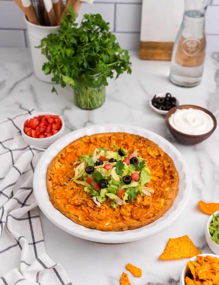 Fully baked taco pie in a white pie dish. The top is golden brown with crispy crushed tortilla chips, resting on a marble countertop with a checkered kitchen towel beside it.