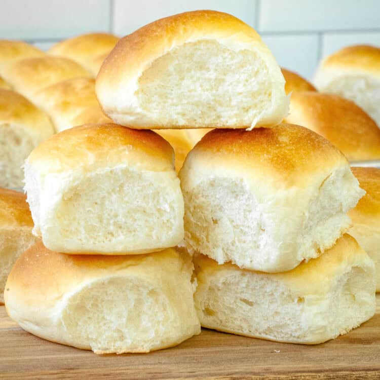 A close-up of freshly baked dinner rolls with a golden-brown top and a soft, fluffy interior. The rolls are stacked on a wooden surface, with more rolls in the background, creating a warm and inviting homemade feel.
