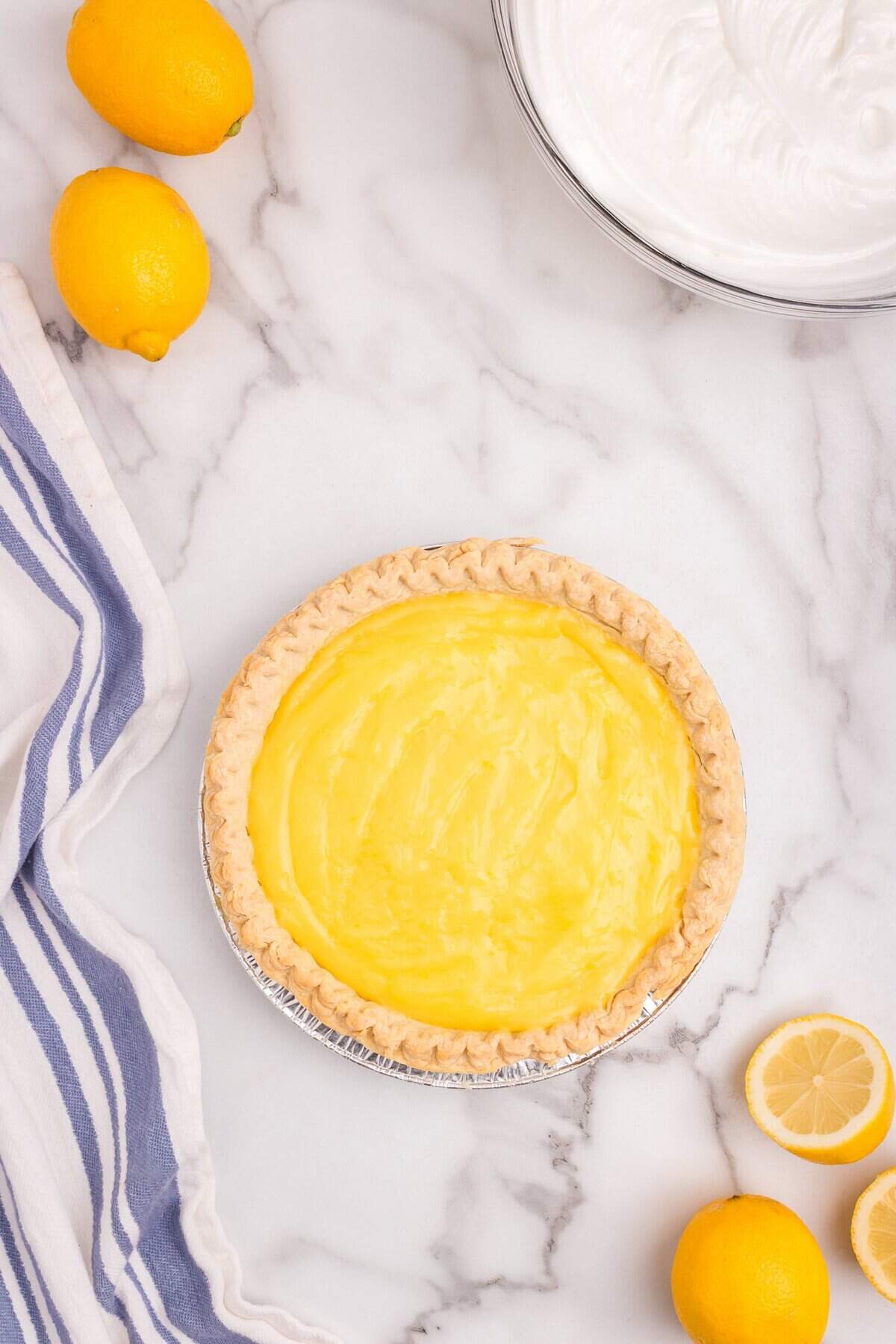 Lemon custard spread evenly inside a baked pie crust, with lemons and a bowl of meringue in the background.