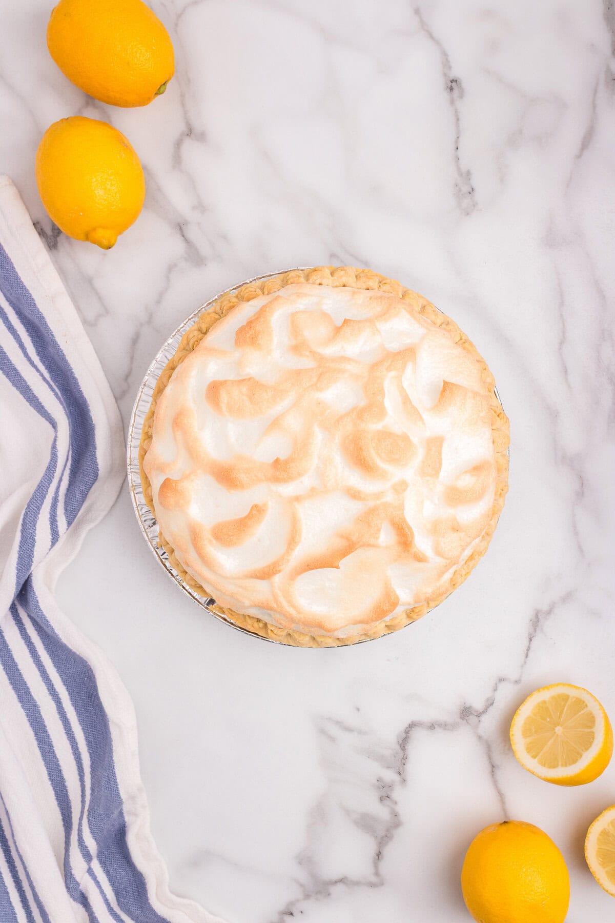 Fully baked lemon meringue pie with golden-brown meringue peaks, sitting on a marble countertop with lemons nearby.