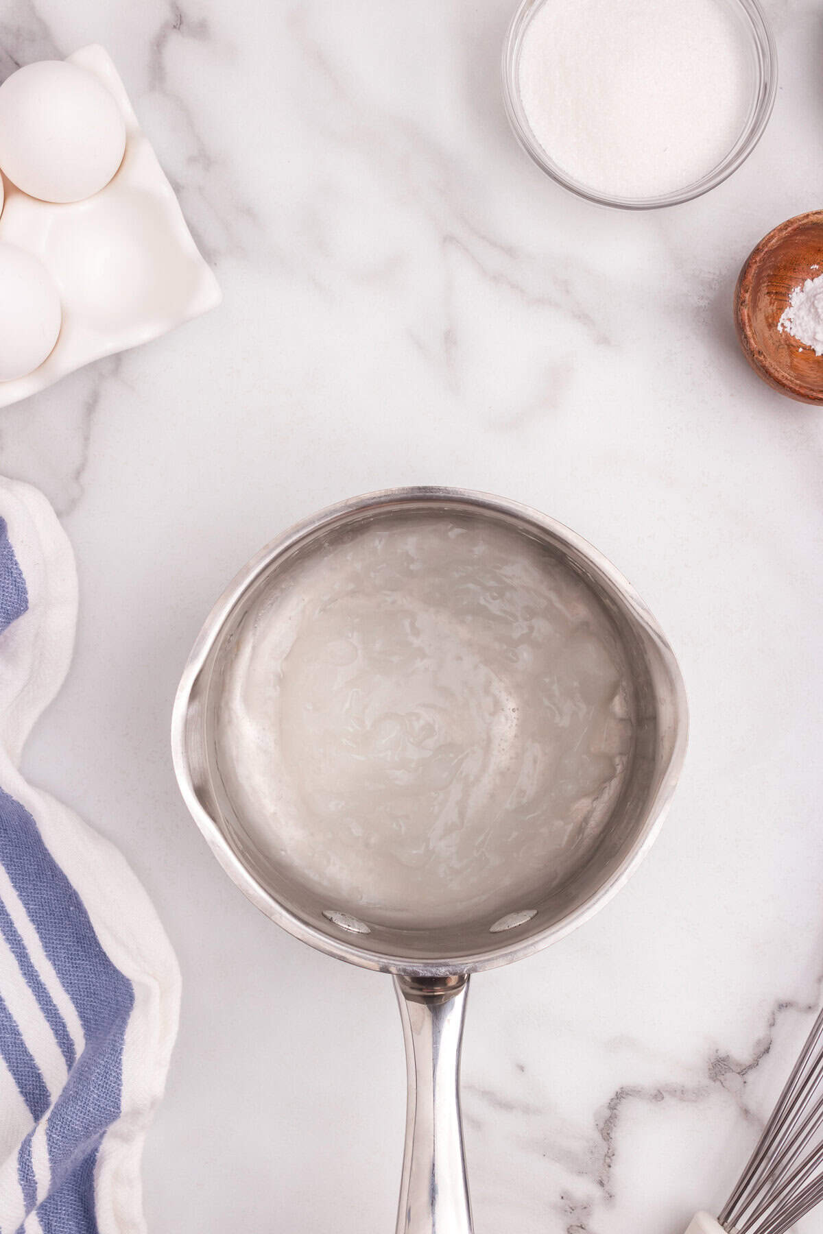 A saucepan with a translucent cornstarch and water mixture thickened, sitting on a marble surface with eggs, sugar, and salt nearby.