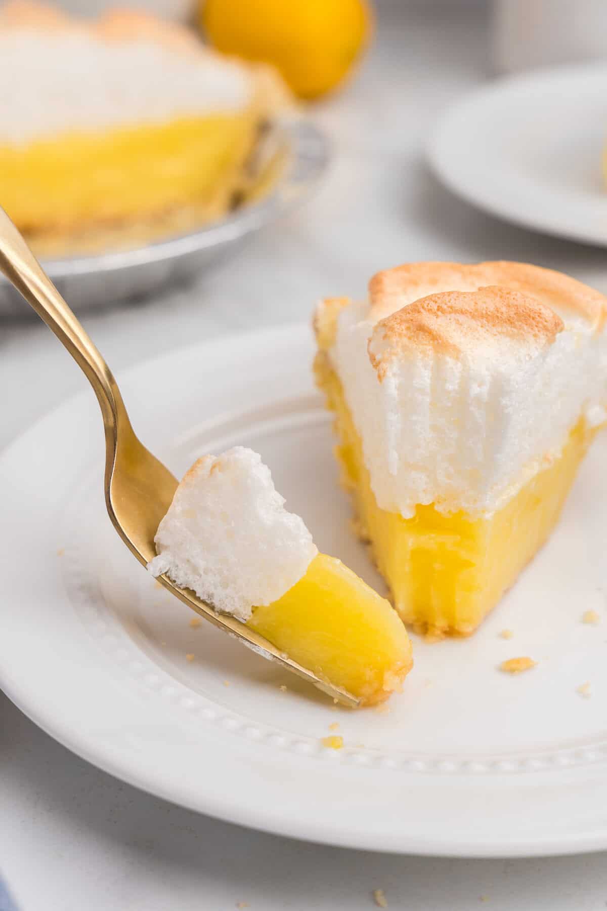 A single slice of lemon meringue pie served on a white plate with a gold fork, showing the smooth lemon filling and thick meringue topping.
