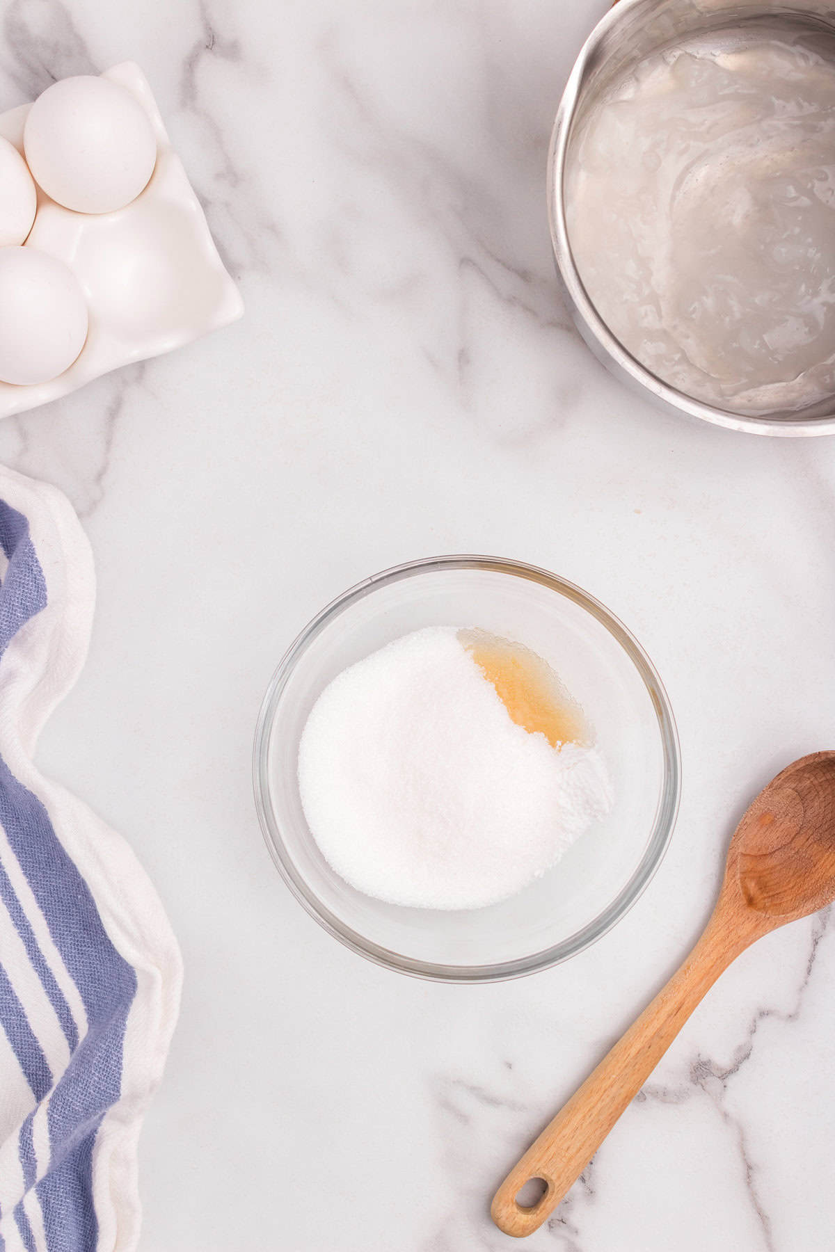 A glass bowl with sugar and vanilla extract placed on a counter, with the saucepan of thickened mixture in the background.