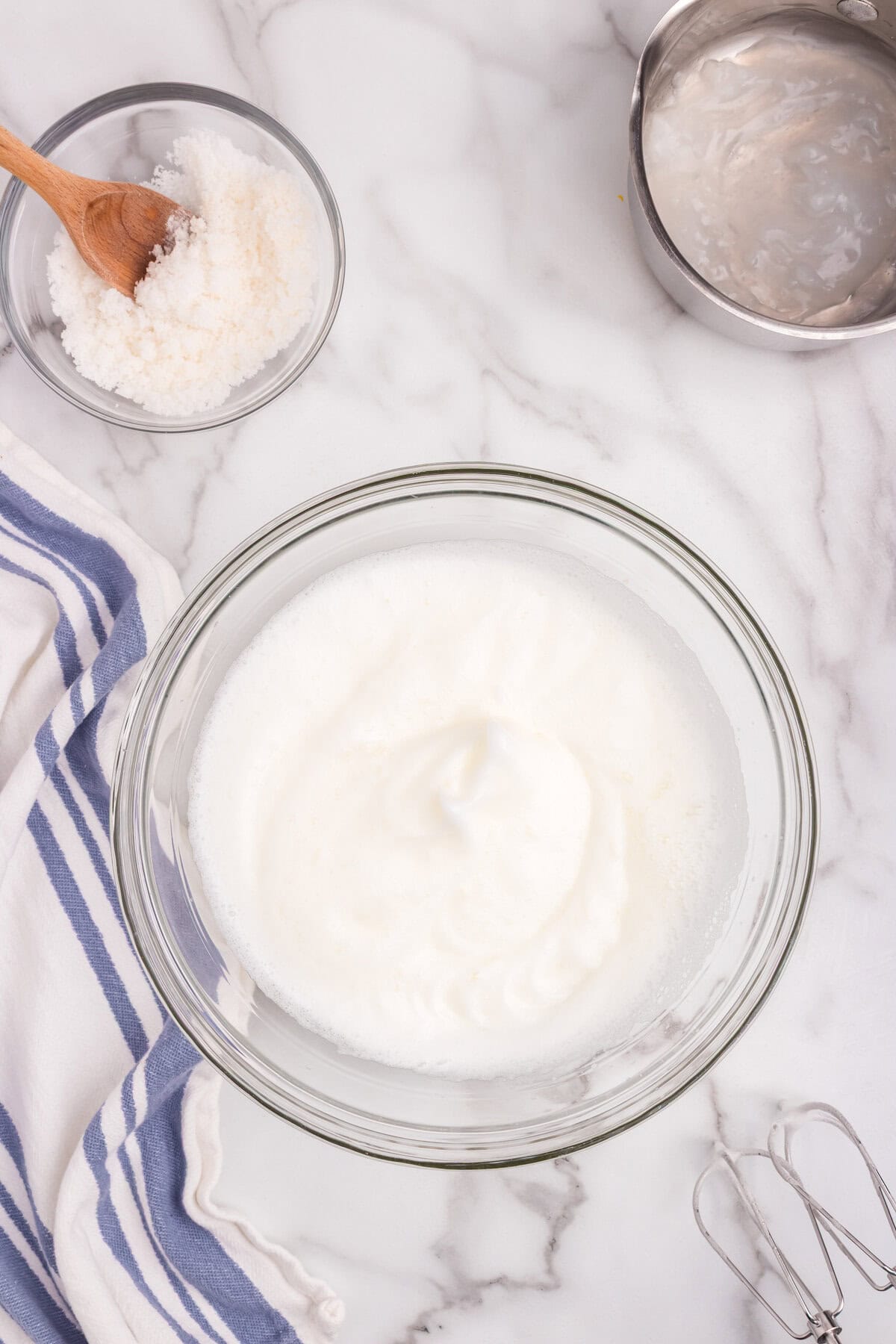 A bowl of whipped egg whites beaten to soft peaks, with a small dish of sugar mixture and the saucepan of cornstarch mixture nearby.