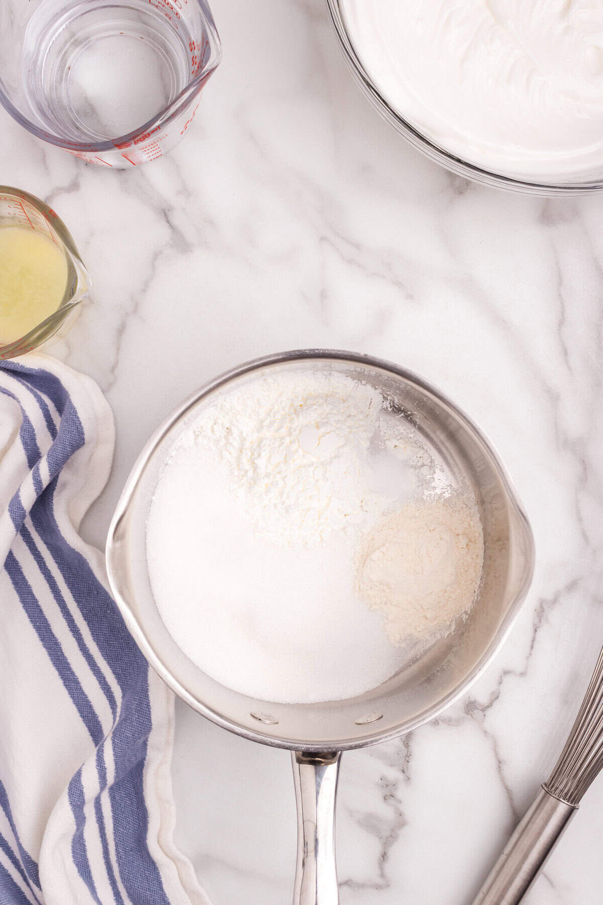 A saucepan filled with sugar and flour before cooking, with measuring cups of water and lemon juice beside it.