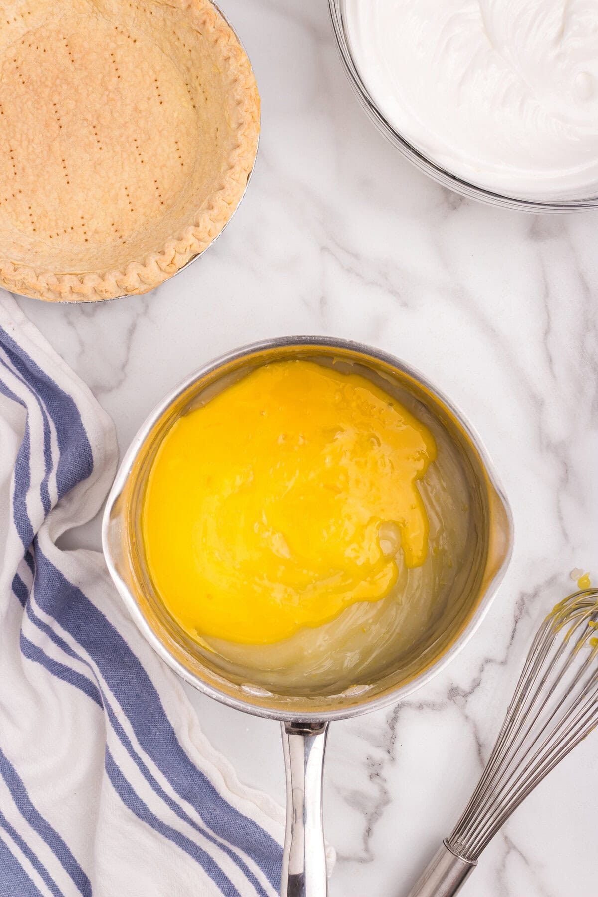 A saucepan with lemon custard thickening to a bright yellow, a baked pie crust set beside it.