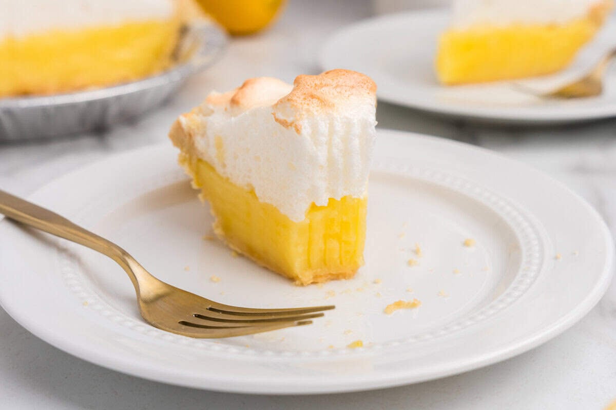 Close-up of a fork cutting into a slice of lemon meringue pie, highlighting the layers of bright yellow lemon curd and fluffy toasted meringue.