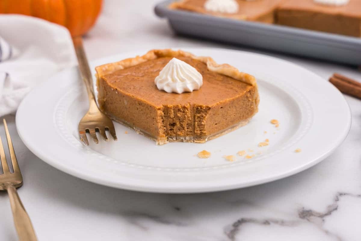 Slice of pumpkin pie bar with a bite taken out, topped with a swirl of whipped cream, served on a white plate with a fork.