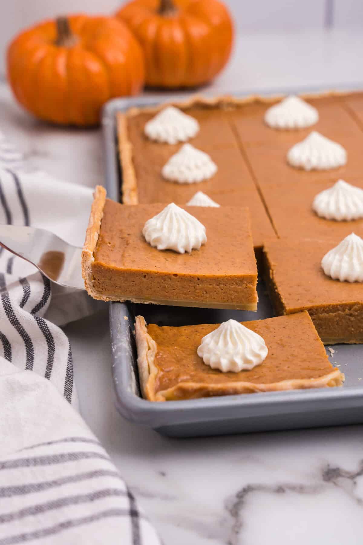Close-up of a pumpkin pie bar being lifted from a baking pan, topped with a swirl of whipped cream, with more bars in the background.