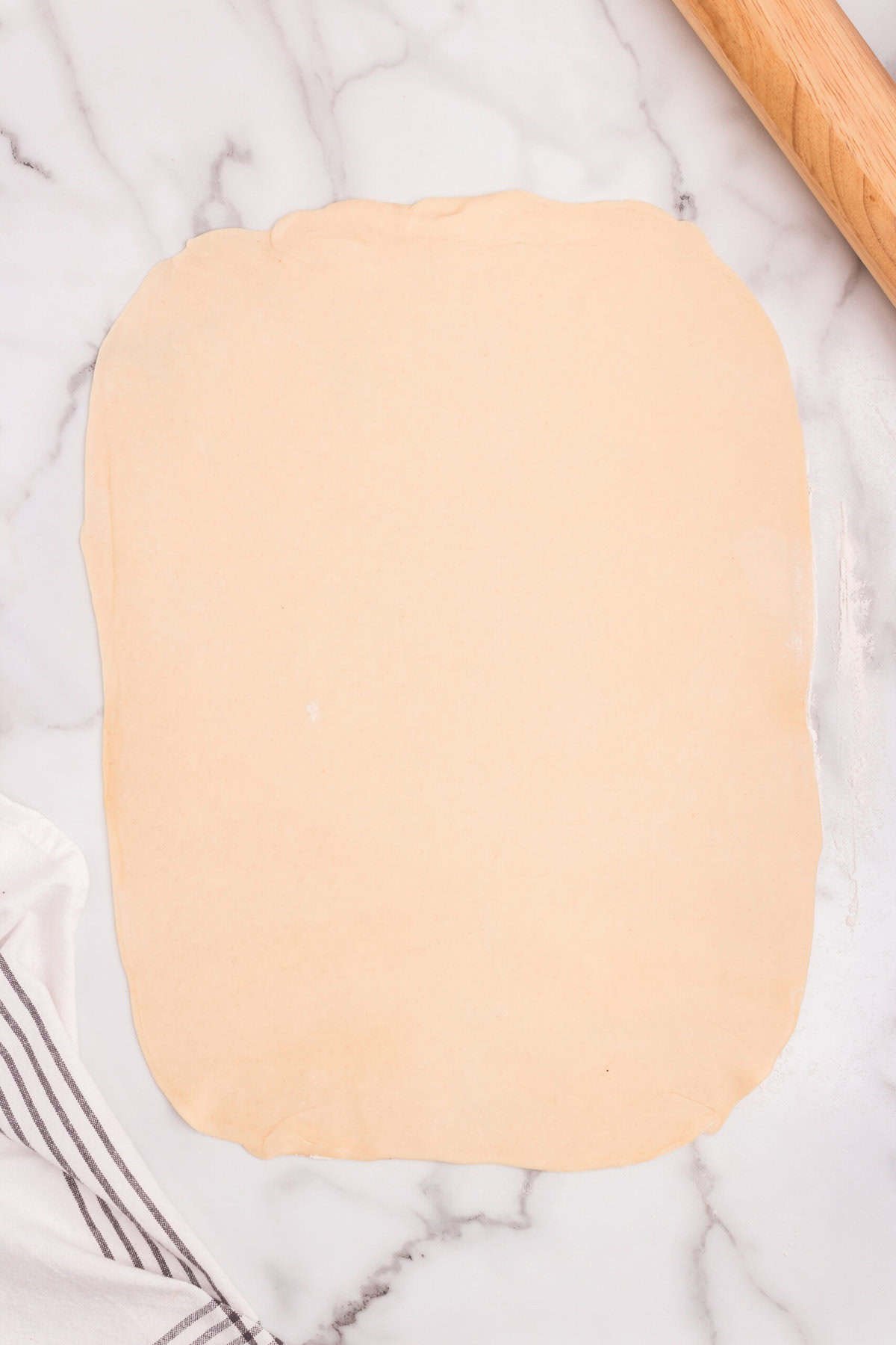 Pie dough rolled into a large rectangular shape on a marble surface with a rolling pin nearby.